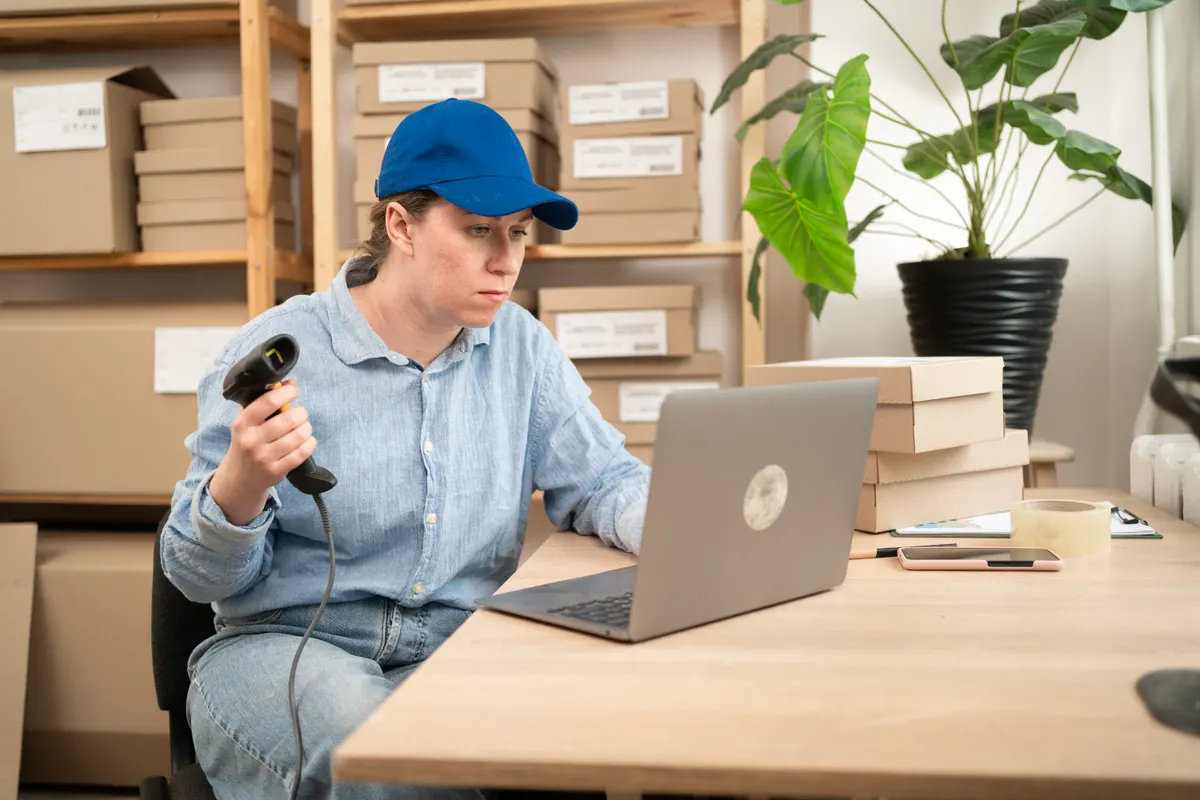 A store owner scanning a product at her laptop, surrounded by boxes and plants