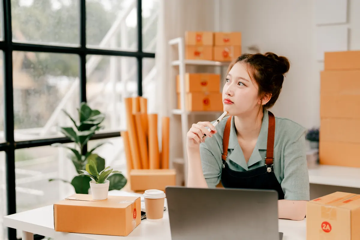 Average order value playbook: young asian woman entrepreneur working on e-commerce business at home, using laptop and organizing cardboard shipping boxes, contemplating new strategies and growth for her online store.