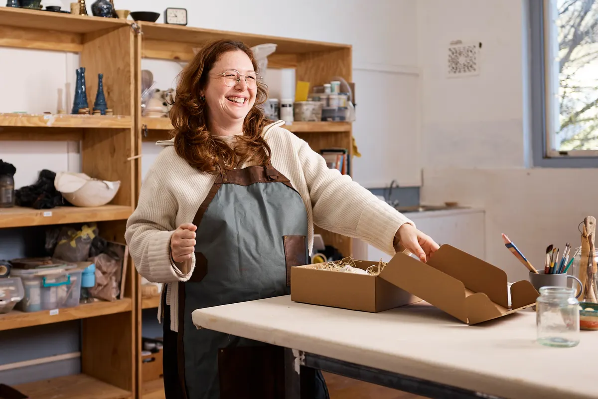 Bundling and upsell: smiling Pottery Studio Owner Packing Handmade Ceramics in Cardboard Box in Creative Workshop.