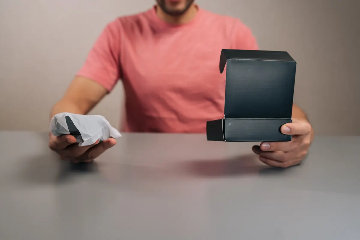 Post-purchase flow: detail cropped shot of person unboxing new electronic gadget, removing from black retail packaging on gray table, signifying new purchase, consumerism, technology.