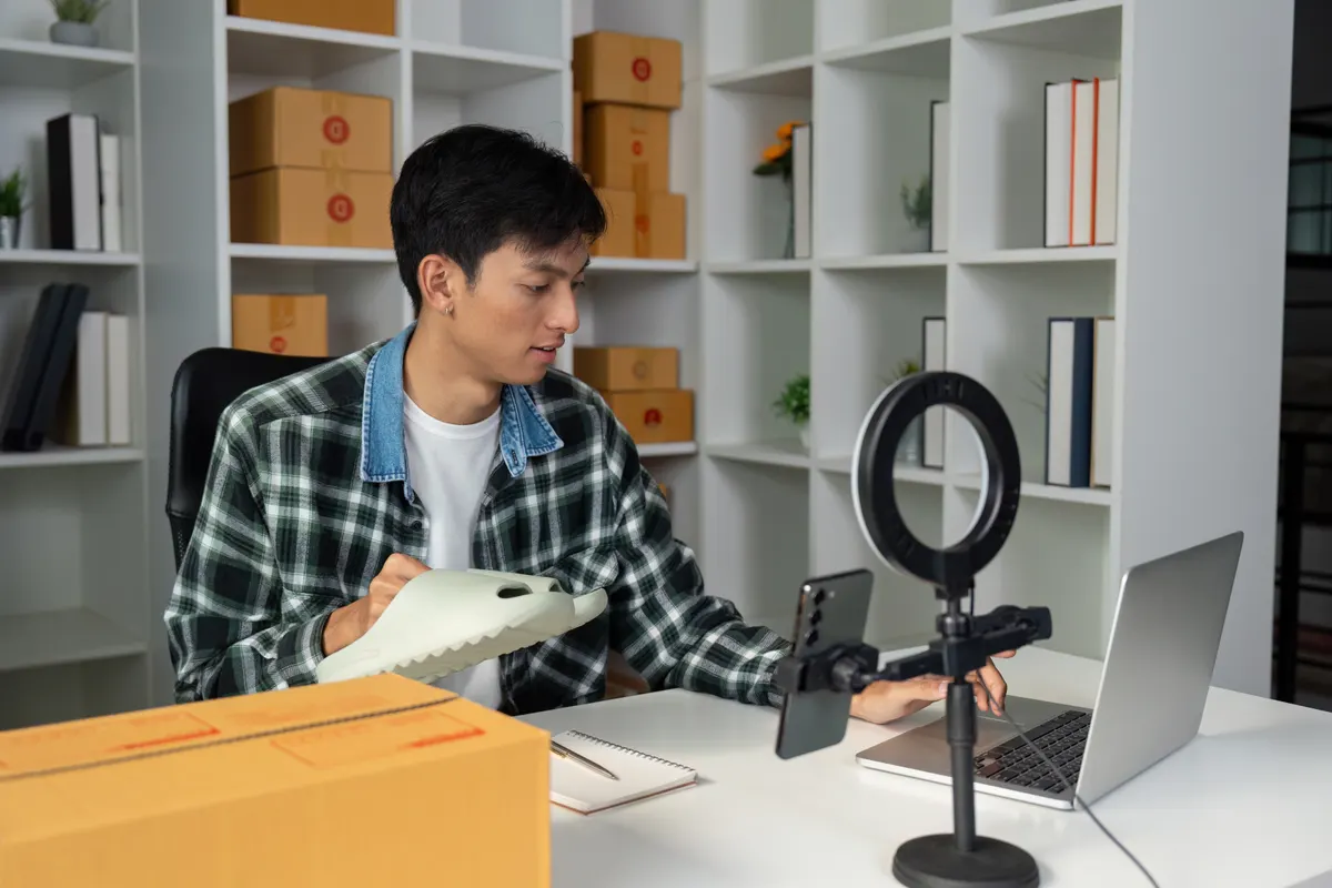 Product feed optimization: young entrepreneur managing online sales while showcasing product, using laptop and smartphone in modern workspace.