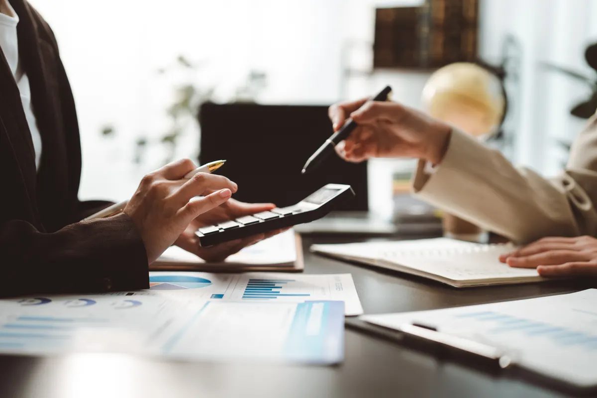 ROAS, MER and CAC explained: financial professionals collaborating on business project. One person calculating data with a calculator. Another person pointing at documents. Analyzing company revenue. Managing finance.