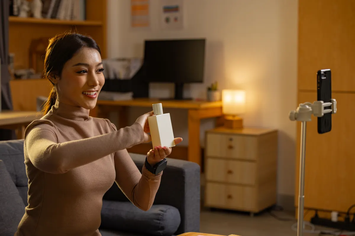 UGC for ecommerce ads: happy asian influencer woman recording live video stream holding cosmetic bottle with cheerful smile at home night time using smartphone on tripod for social media review showing beauty product.