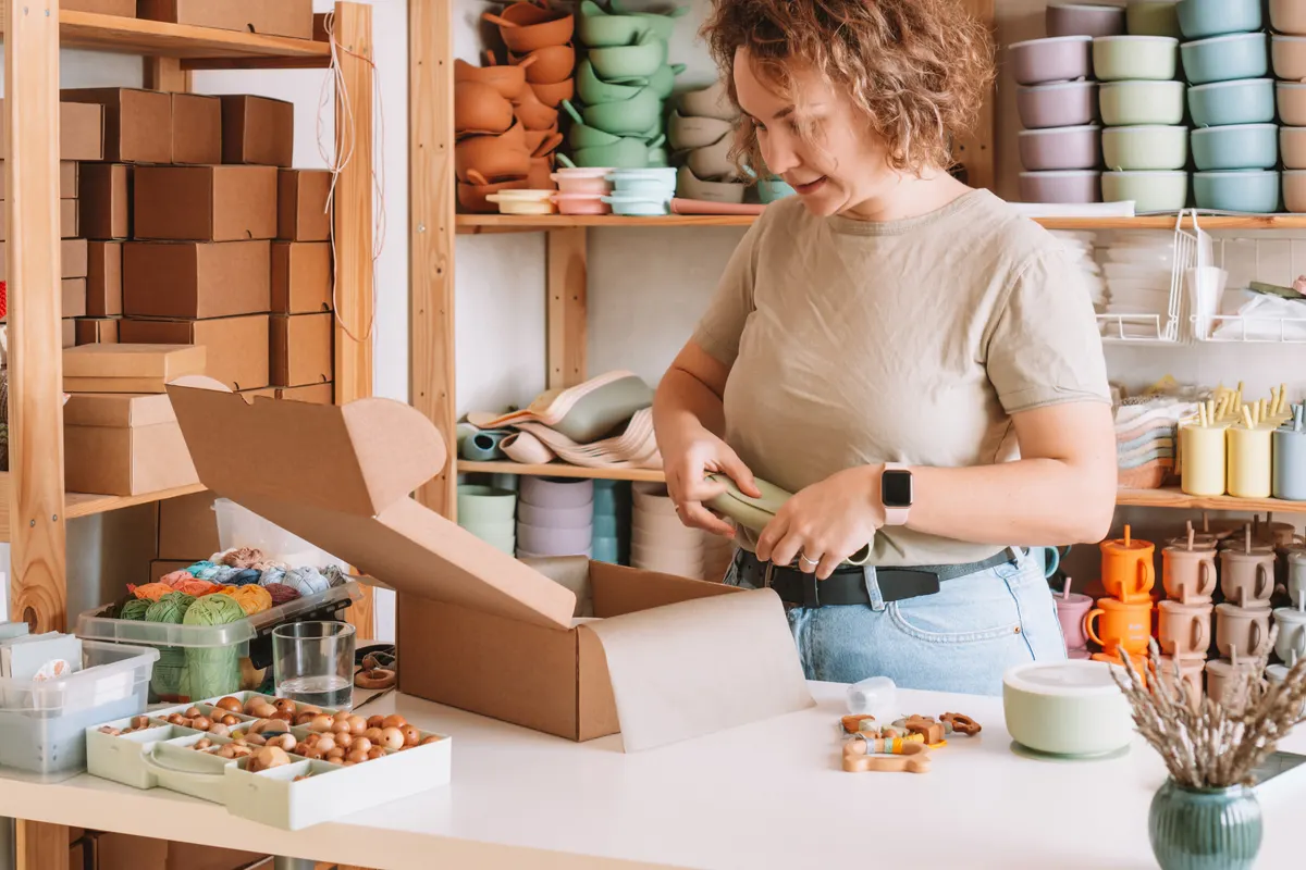 Toy ecommerce marketing agency: smiling concentrated curly woman packing and wrapping shipment with silicone baby dish in box for delivery. Work at home.