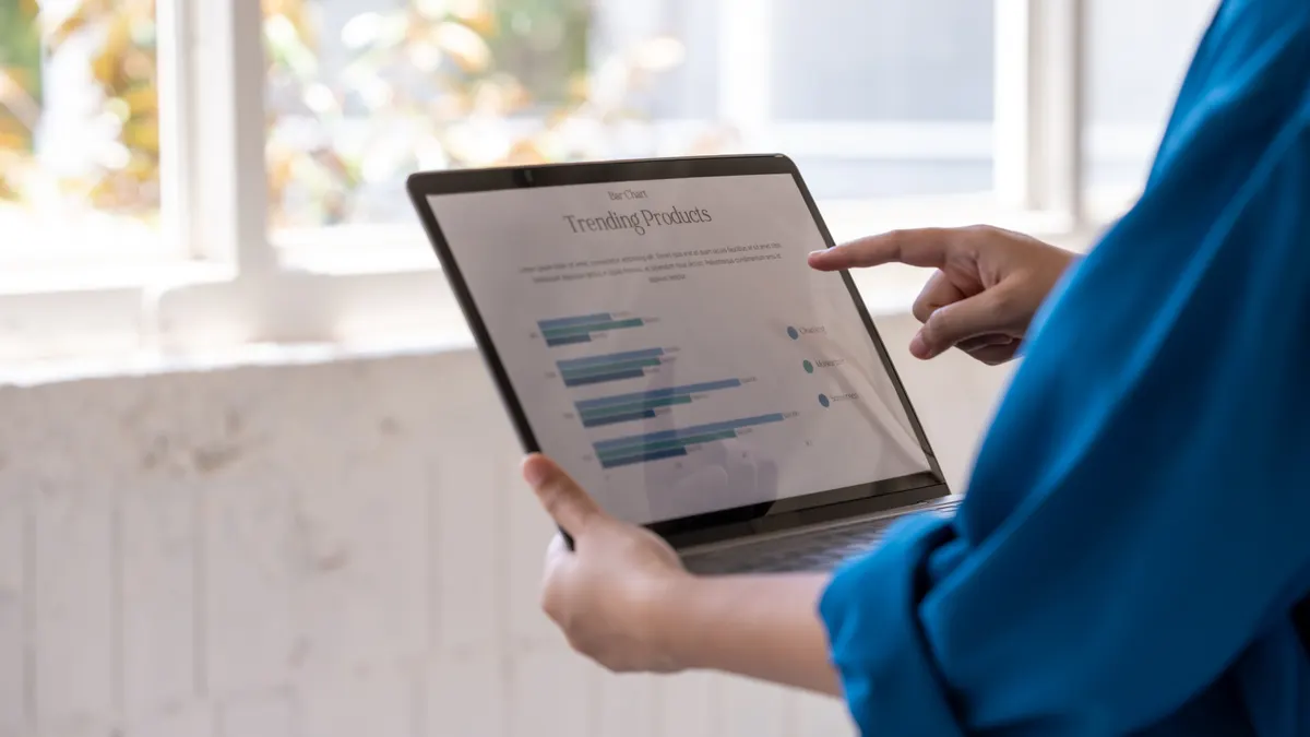 How to read a Shopify analytics dashboard: close view of a person in a blue shirt holding a laptop with charts on screen near a bright window, showing business data, remote work, analysis, and modern office use.