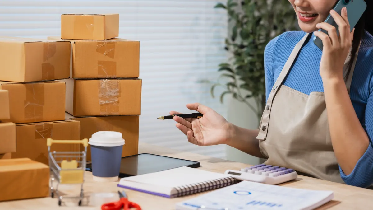 How to set ad budgets by margin, not revenue: small business owner packing online orders at a desk, talking on the phone beside stacked cardboard boxes, calculator, notebook and coffee cup in a home office.
