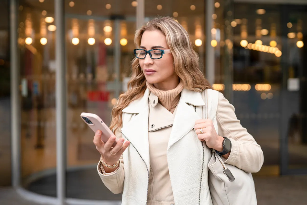 SMS consent done properly: woman stands outside a building looking at her smartphone while holding a bag.