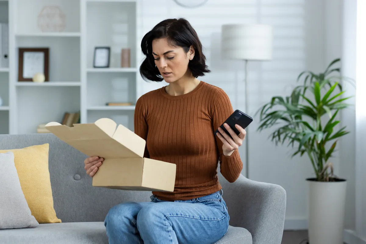The post-purchase email people actually open: an upset young woman is sitting on the sofa at home, holding a mobile phone in her hand and worriedly looking into an open box of a parcel that she received by mistake.