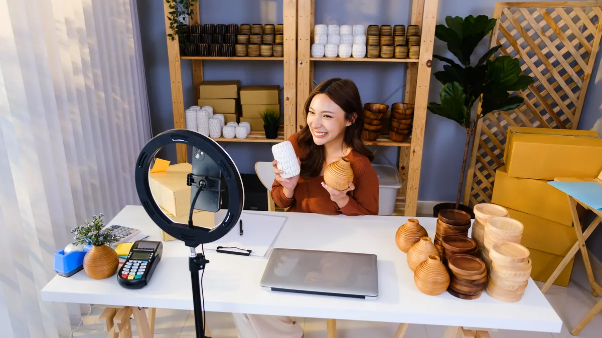 What changed in Google shopping this year: smiling young woman hosting a live stream in a home studio, showcasing handcrafted vases and bowls for online sale, promoting her small business in modern ecommerce and digital marketing style.
