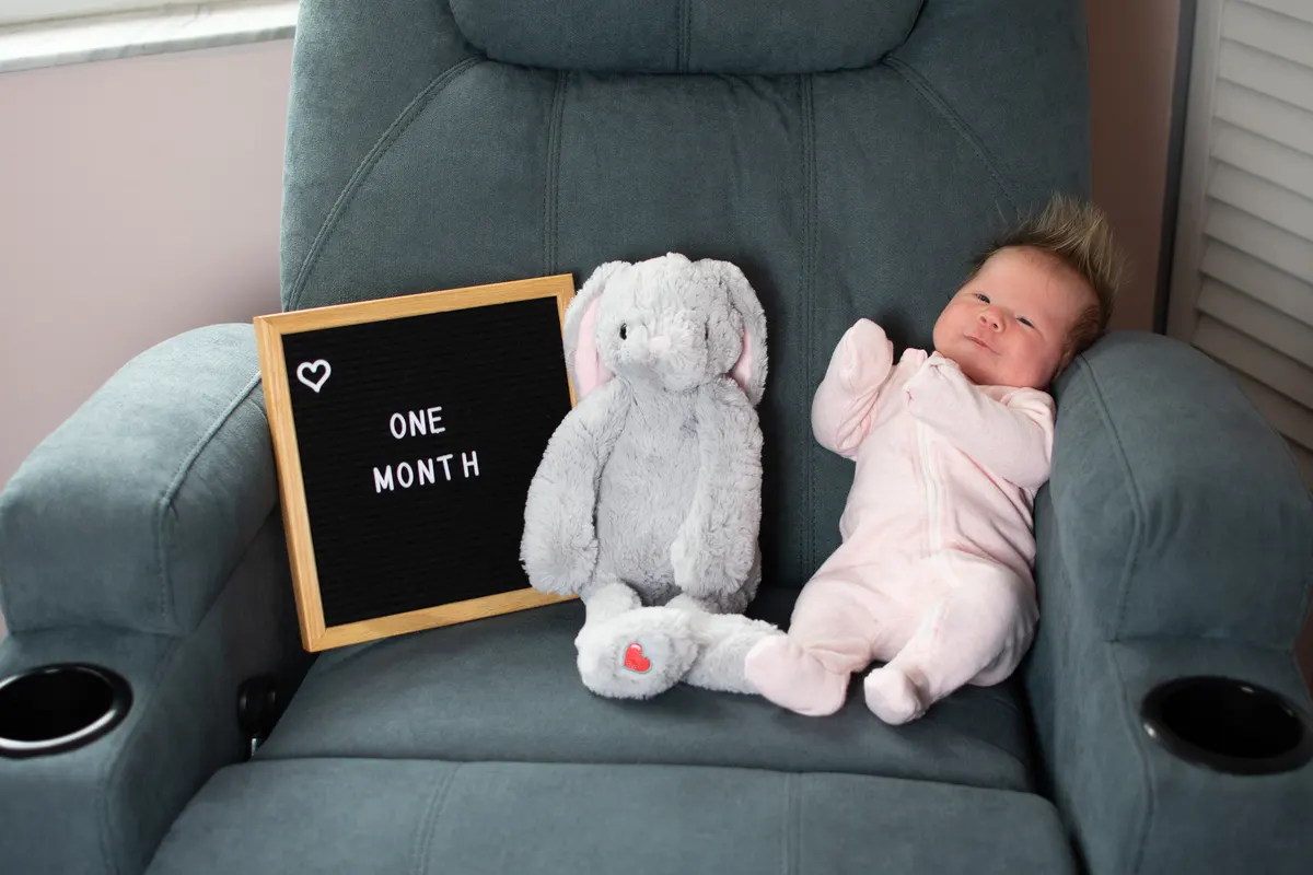 What to expect in the first month with us: one month old Caucasian baby girl near the plush toy and letter board. Newborn baby monthly picture.