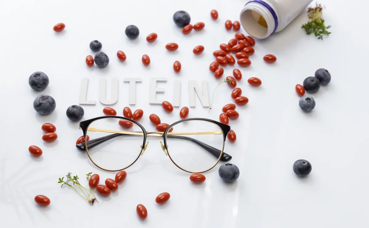 Why supplement brands get disapproved: lutein capsules scattered from open bottle with pair of eyeglasses fresh blueberries green plant leaves and text lutein on clean white background concept.