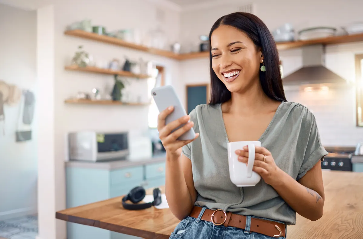 A woman smiling at her phone in a bright kitchen, browsing a store
