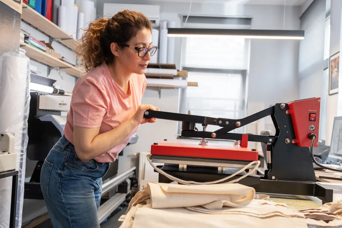 Print-on-demand apparel marketing agency: a print shop worker is operating a heat press machine for textile printing, carefully aligning the material before pressing.