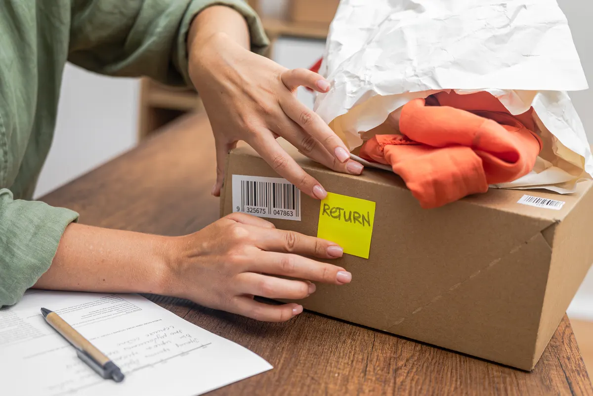 Wholesale-to-trade store marketing agency: woman online shopper affixes a barcode sticker to a cardboard box, marking it for return and refund.