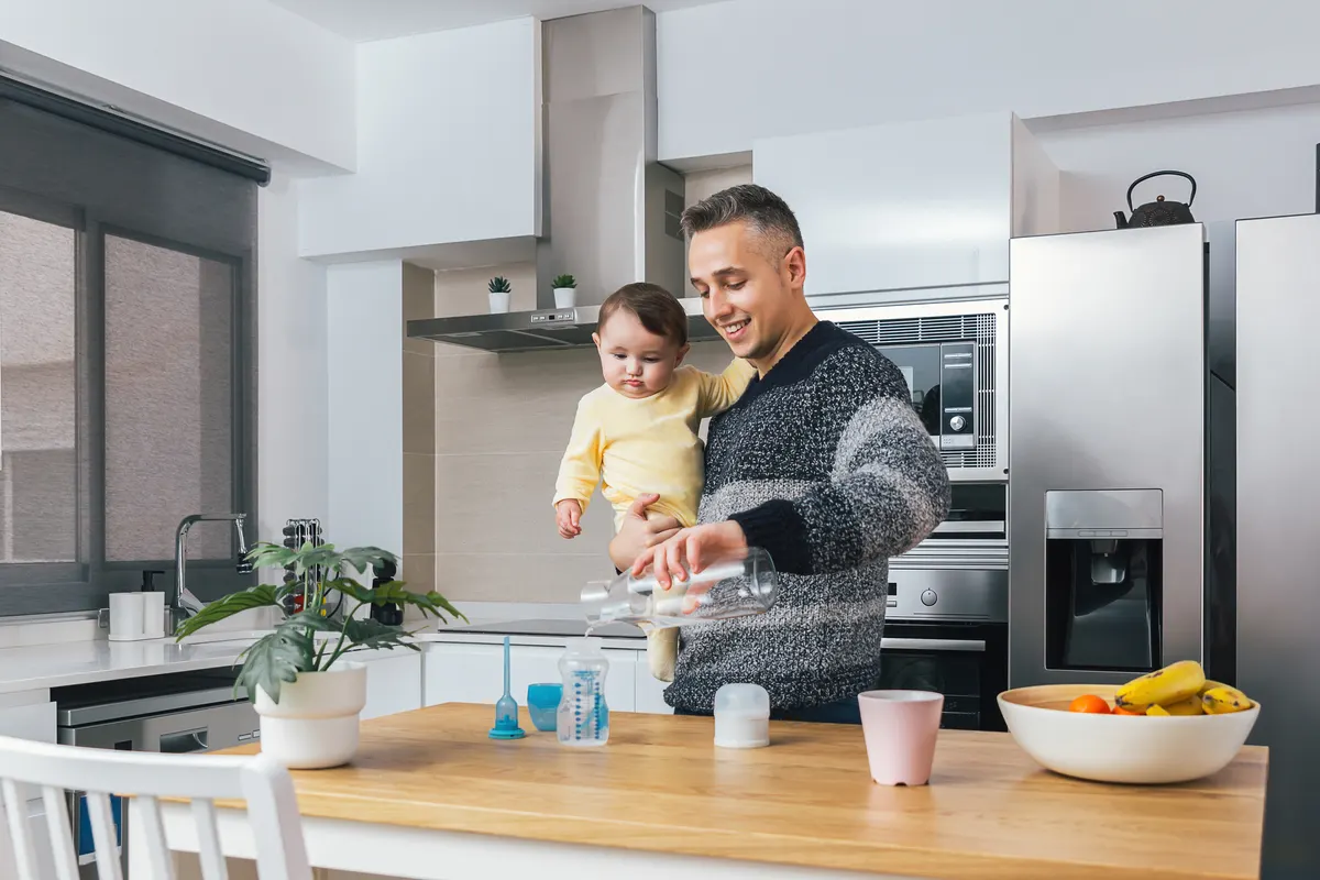 Baby feeding product marketing agency: young single father preparing a feeding bottle while holding his baby in arms in the kitchen. Man and child, parenting fatherhood real moment. Single dad family home life concept.