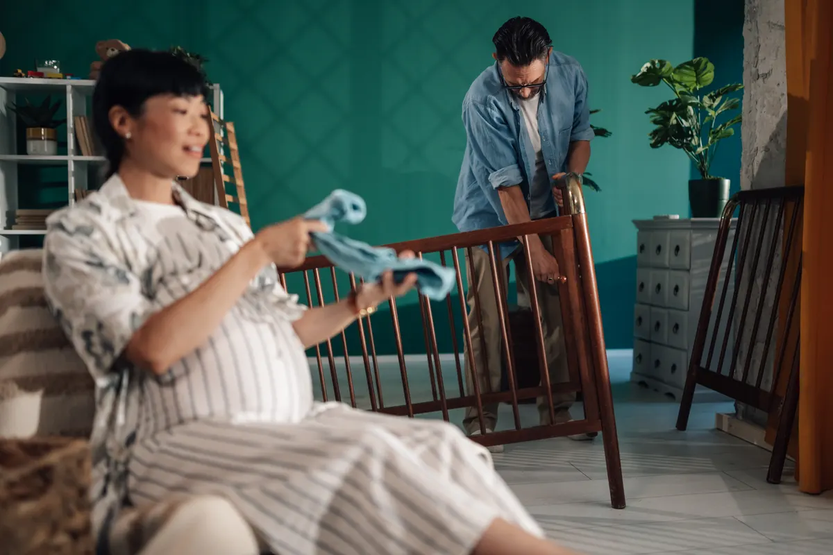 Nursery furniture marketing agency: pregnant woman holding baby clothes while man assembling crib.