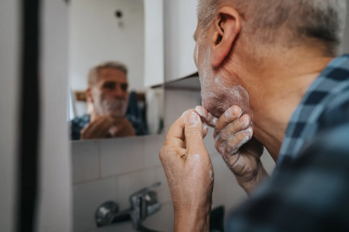Men's grooming marketing agency: senior man shaving at bathroom mirror, focusing on grooming routine and reflection during morning routine.
