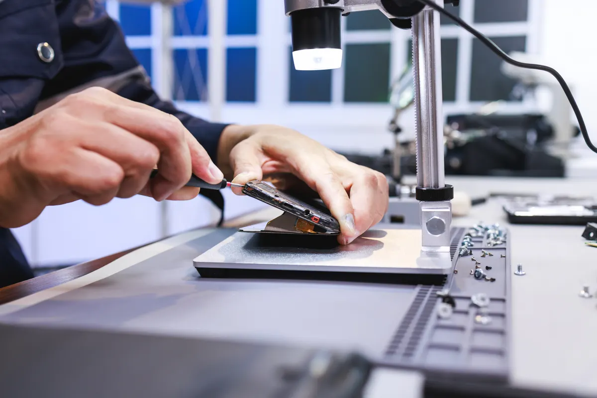 Refurbished electronics marketing agency: close-up of a technician repairing a smartphone on a workbench under a small lamp, showing phone maintenance, electronics service, and careful hands-on work in a modern workshop.