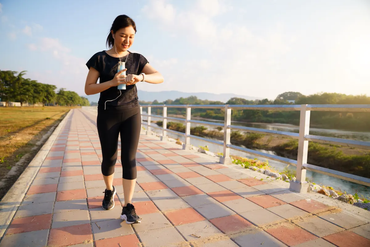 Wearable tech marketing agency: young Asian woman checking her smartwatch while exercising at a riverside park.
