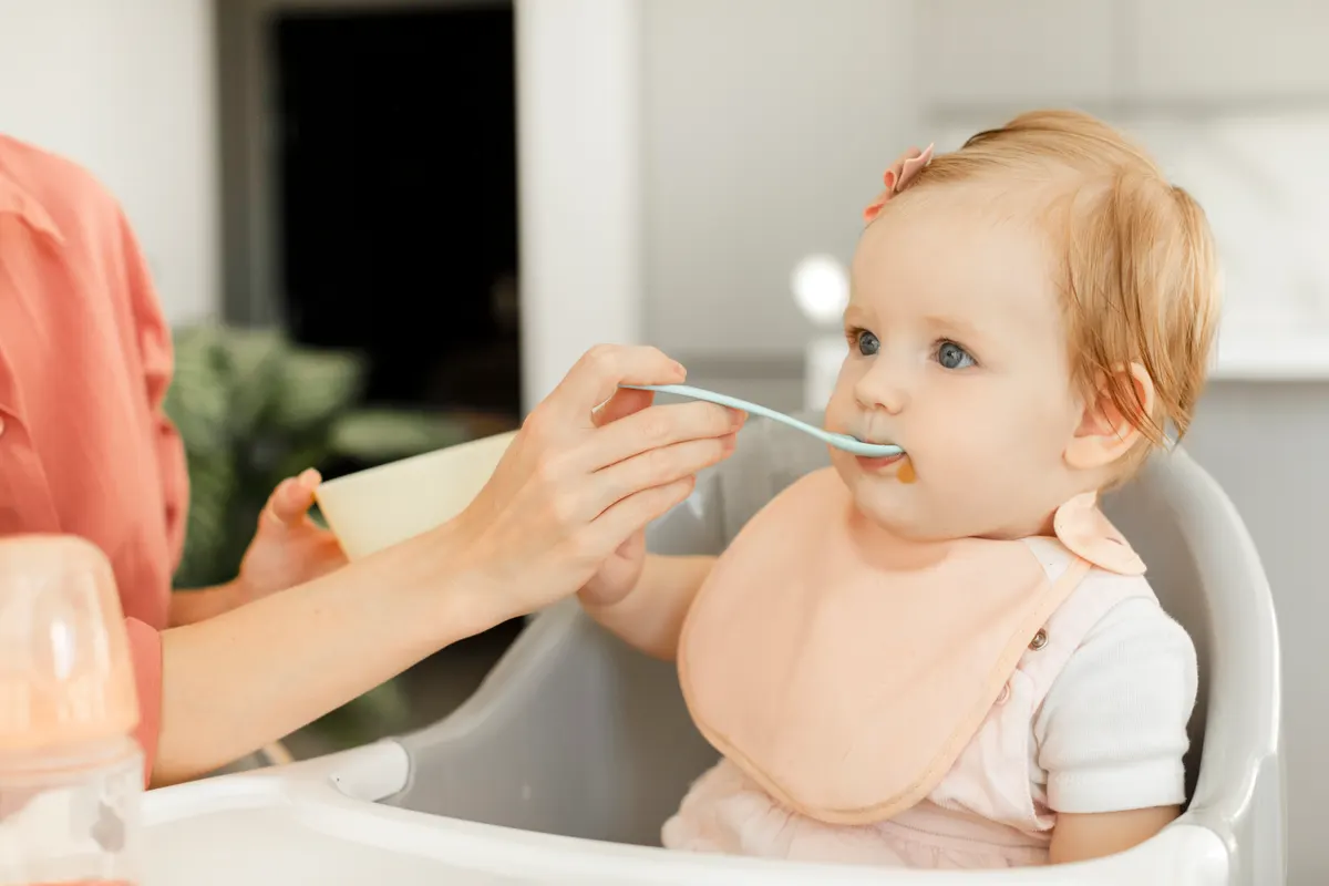 Baby food marketing agency: baby girl eating blend mashed food sitting on high chair while mother feeding child for vegetable lunch. Healthy eating for baby humans.