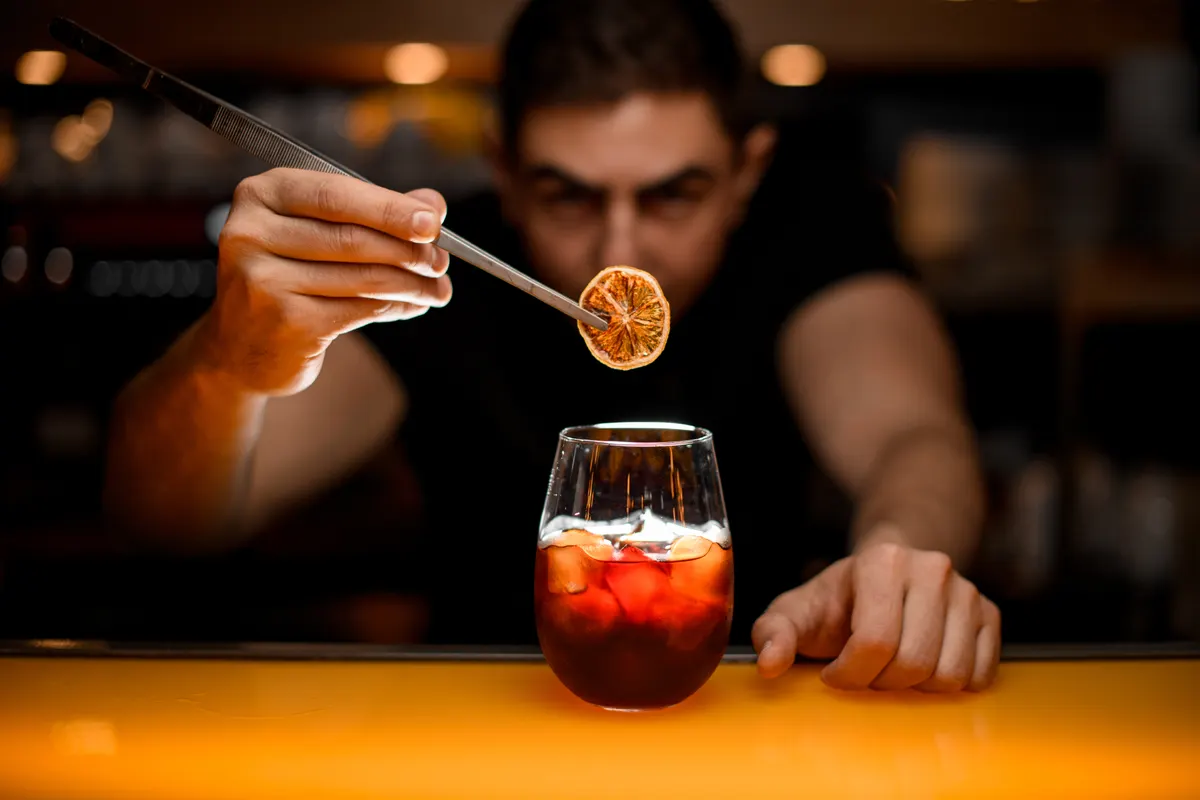 Non-alcoholic drink marketing agency: close-up of glass with cold drink and male hand accurate holding dry slice of orange with tweezers over it.