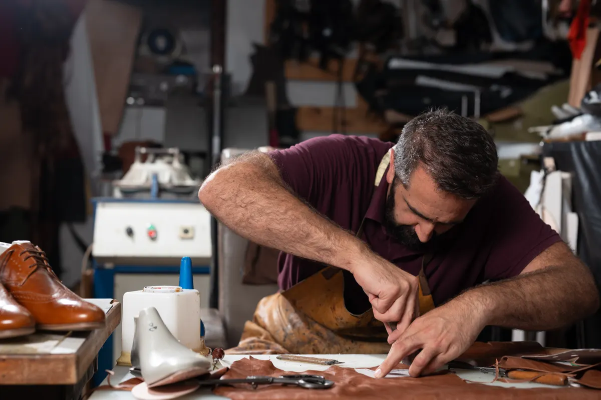 Handmade leather shoe marketing agency: man crafts leather shoes in a cluttered workshop, focused on stitching and detailing.