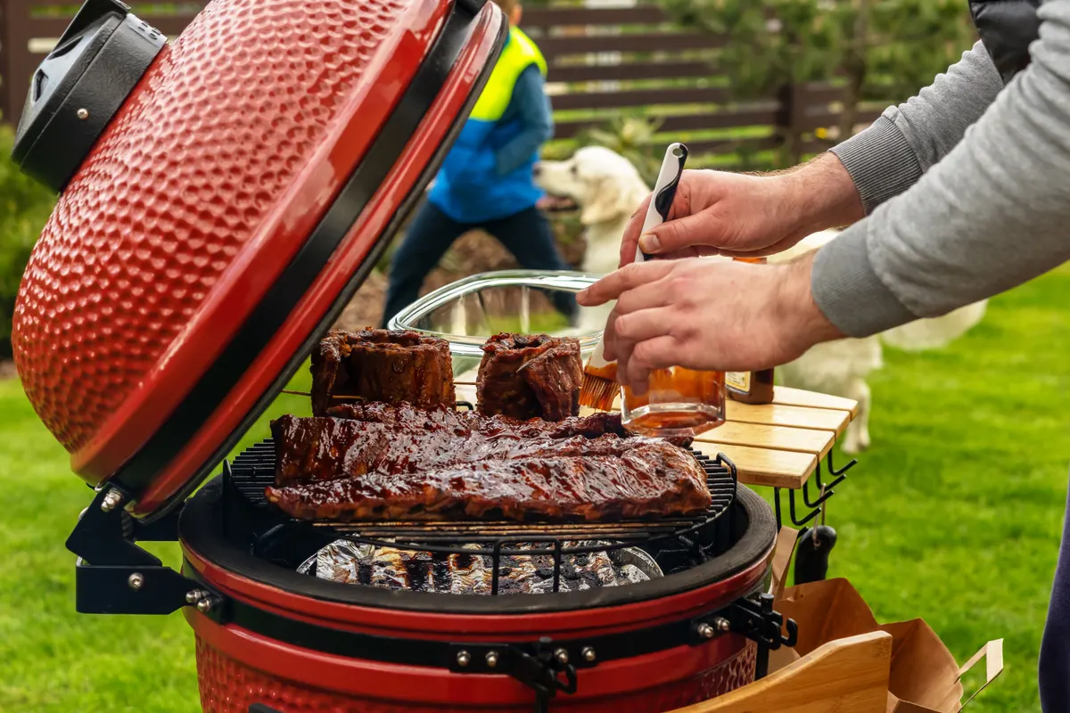 Grill & BBQ marketing agency: red Ceramic Barbecue Grill. The man coats pork ribs with BBQ sauce. Dog and boy playing during family barbecue in the backstage of summer terrace.