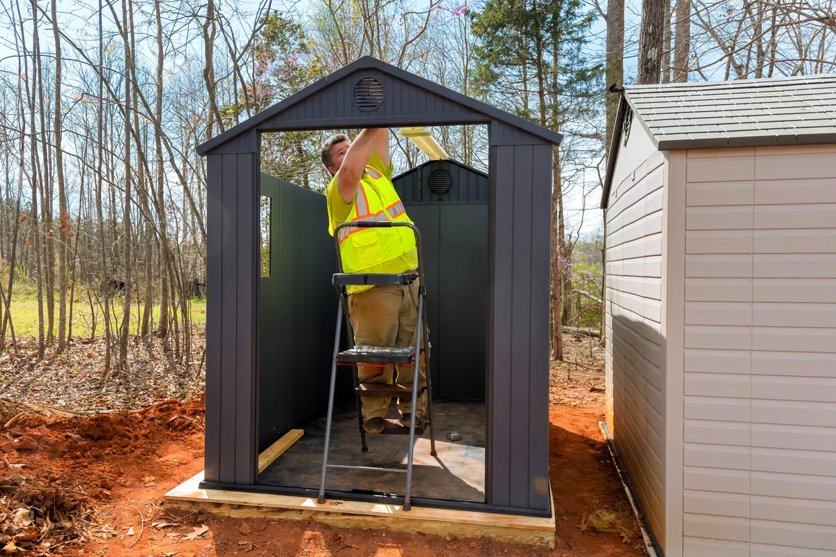 Shed & outdoor structure marketing agency: worker assembling storage shed in clearing surrounded by trees during daylight hours.