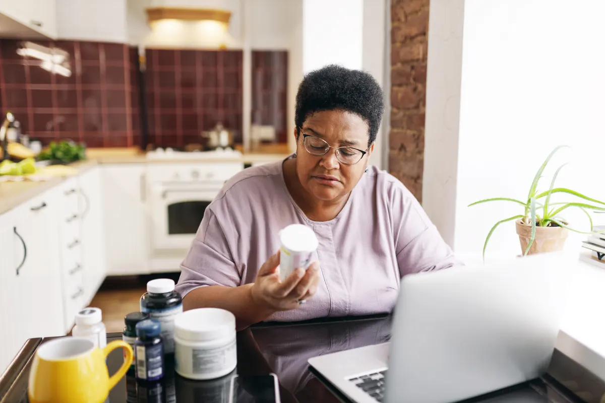 Supplement marketing agency: serious plus size senior black female in glasses sitting at kitchen table full of food supplements using laptop, reading label on vitamin bottle, making online order on marketplace.