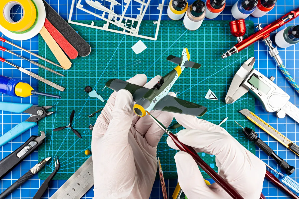 Model kit marketing agency: desktop view from above of assembly and painting of retro scale model fighter plane concept background. modeling tools airbrush gun paint kit parts blue green cutting mat knife and brush work desk.
