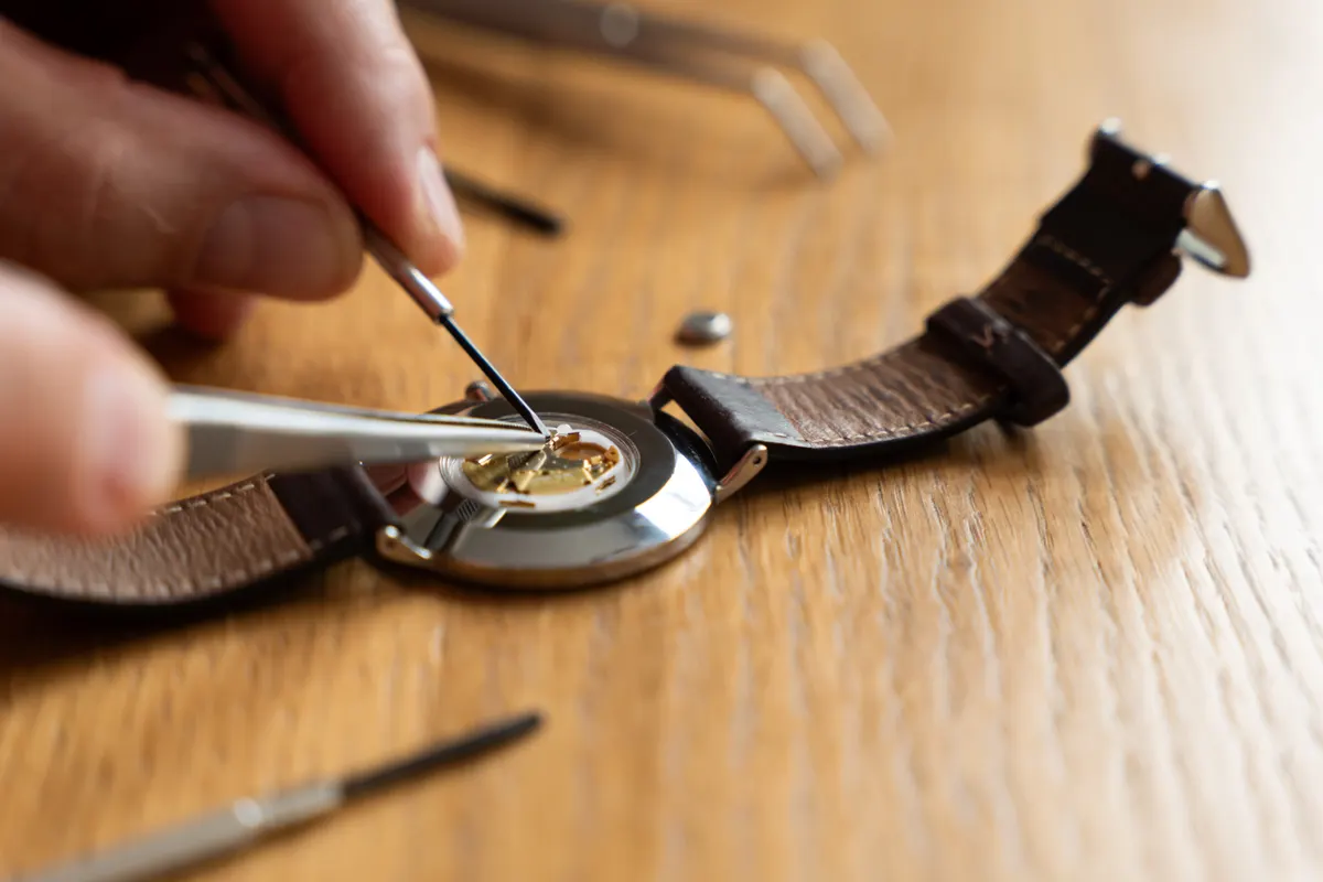 Watch marketing agency: close-up of a person using tweezers to repair a watch movement on a wooden workbench, with tools visible in the background.