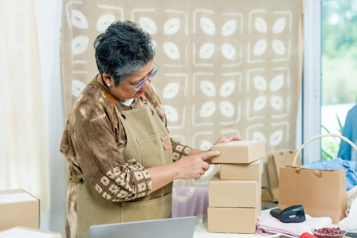 Etsy seller marketing agency: senior asian businesswoman moves packed parcels into neat stack, checks receipt list, prepares courier pickup, home studio craft business order management, calm productivity, concept All set.