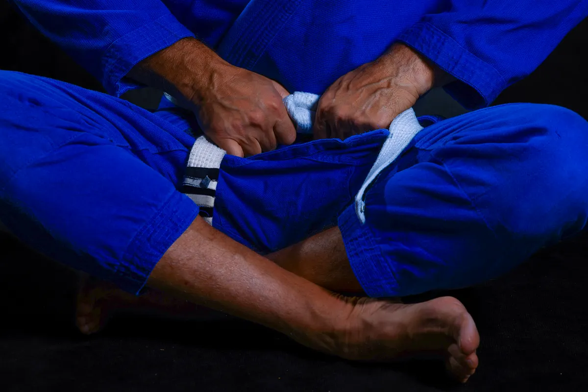 Martial arts gear marketing agency: martial Arts Student Sitting Cross-Legged While Tying White Belt Around Blue Gi, Capturing a Moment of Quiet Preparation, Focused Hands, and the Symbolic Start of a Personal Journey.