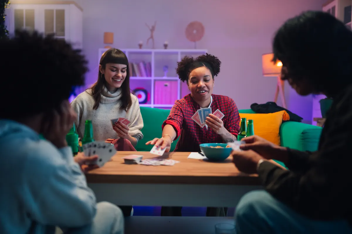 Party game marketing agency: diverse adult friends playing cards and enjoying drinks at a fun game night party in a modern apartment.