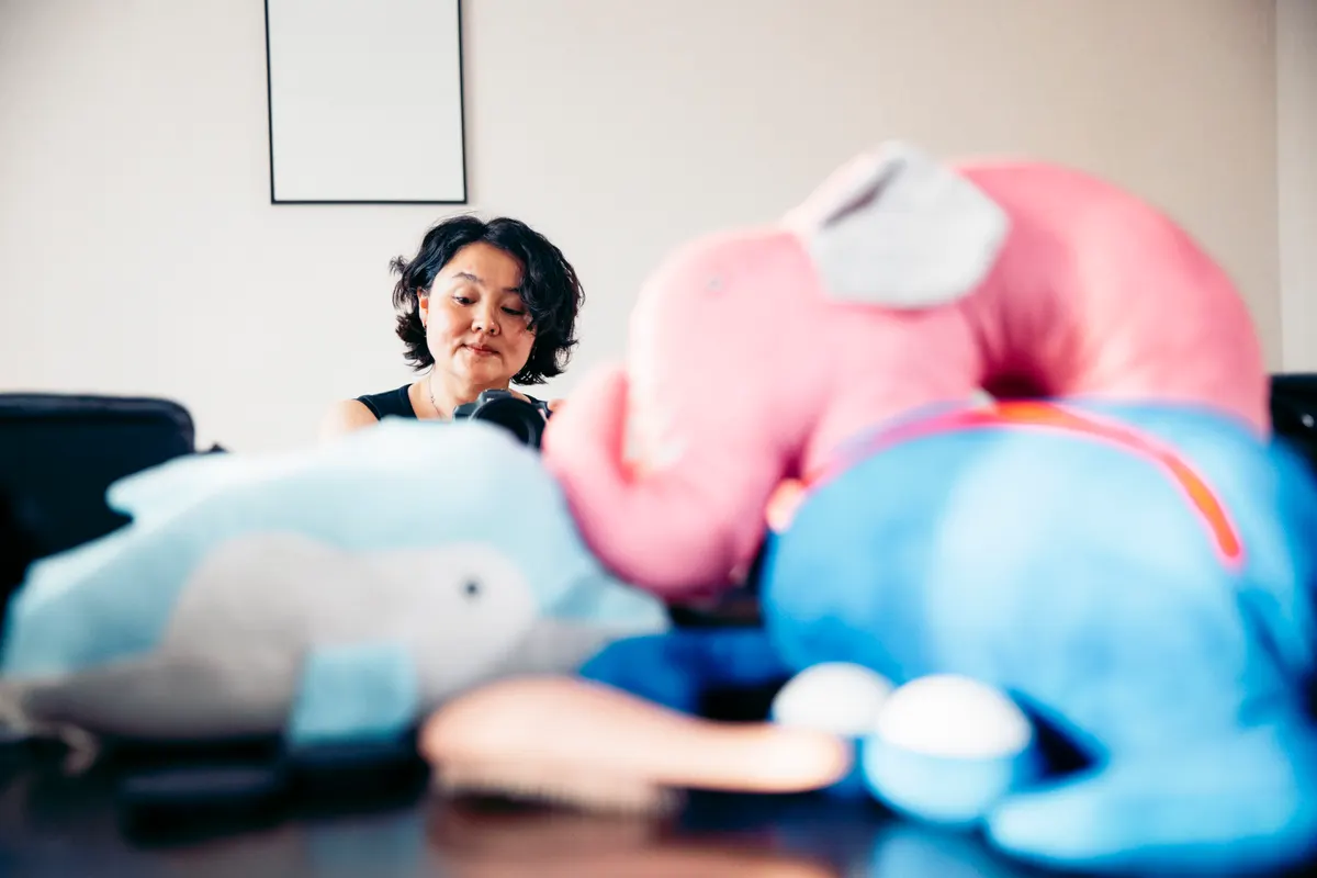 Plush toy marketing agency: asian woman with short curly hair sits at a table surrounded by colorful plush elephant toys. The scene is bright and playful, emphasizing a cozy atmosphere.