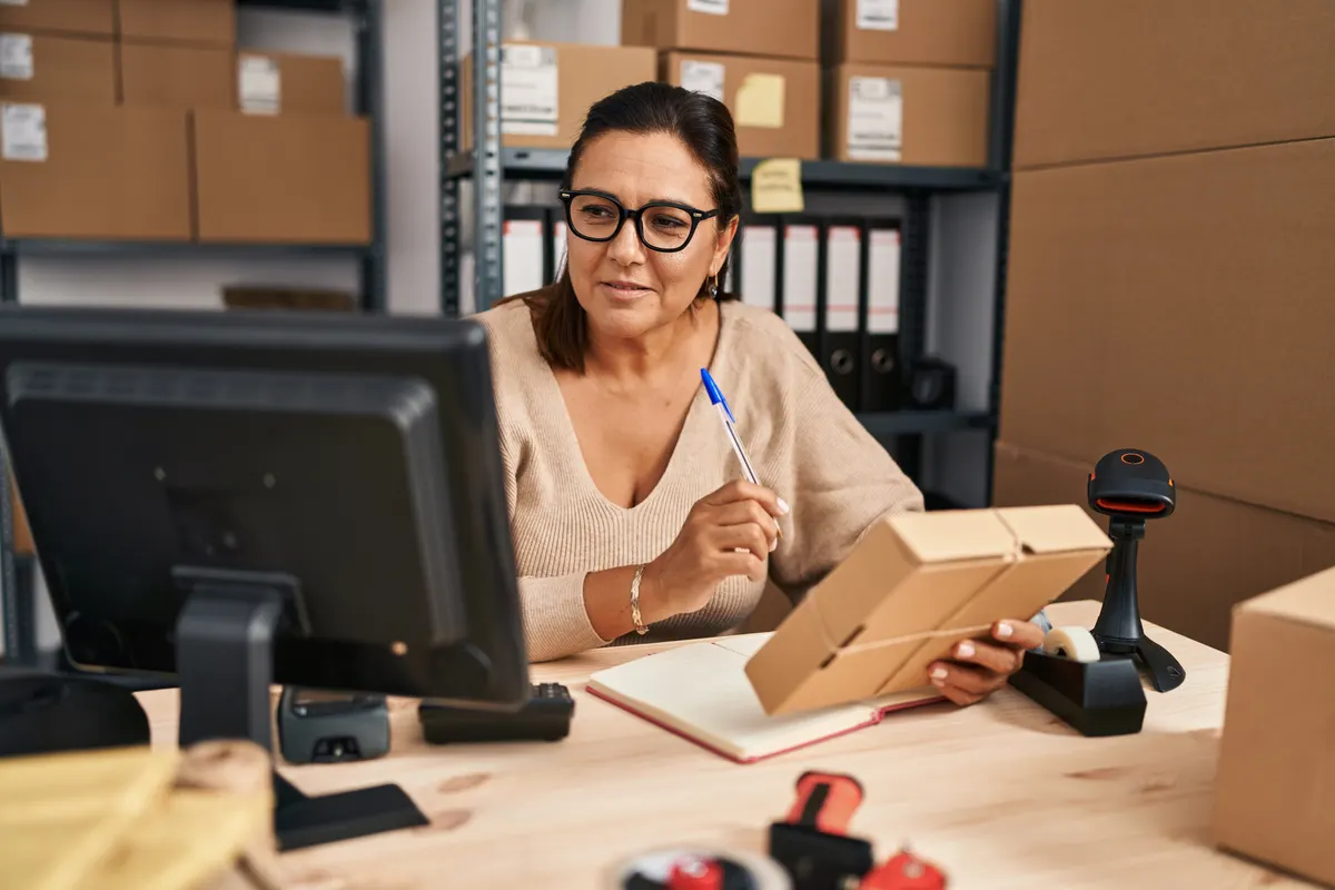 Ecommerce SEO agency: middle age hispanic woman ecommerce business worker writing on notebook holding package at office.